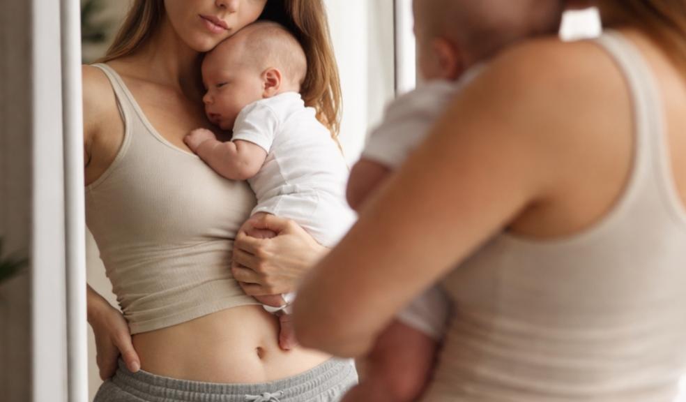 picture of New mum holding baby while looking at her reflection in a mirror representing postpartum body image and motherhood