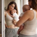 picture of New mum holding baby while looking at her reflection in a mirror representing postpartum body image and motherhood