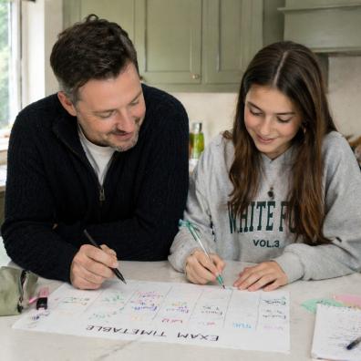 picture of Dad helping his teenage daughter create a revision timetable at a kitchen island in a bright family home