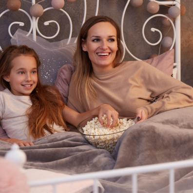 Picture of a mother and daughter snuggled in bed watching a film with popcorn